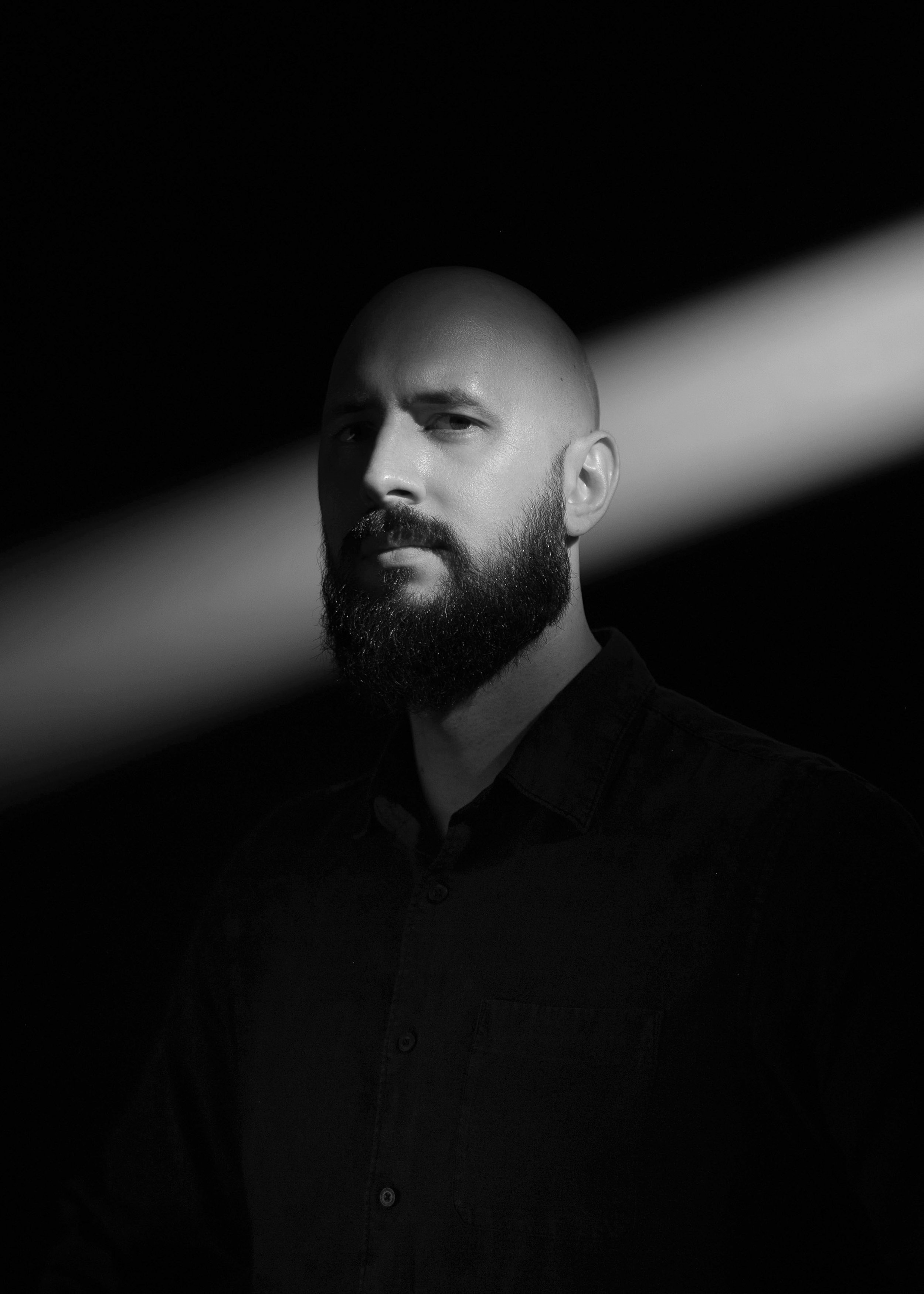 Black and white portrait of a bald man with a beard, looking at the camera with a serious expression, against a dark background with a diagonal light streak.