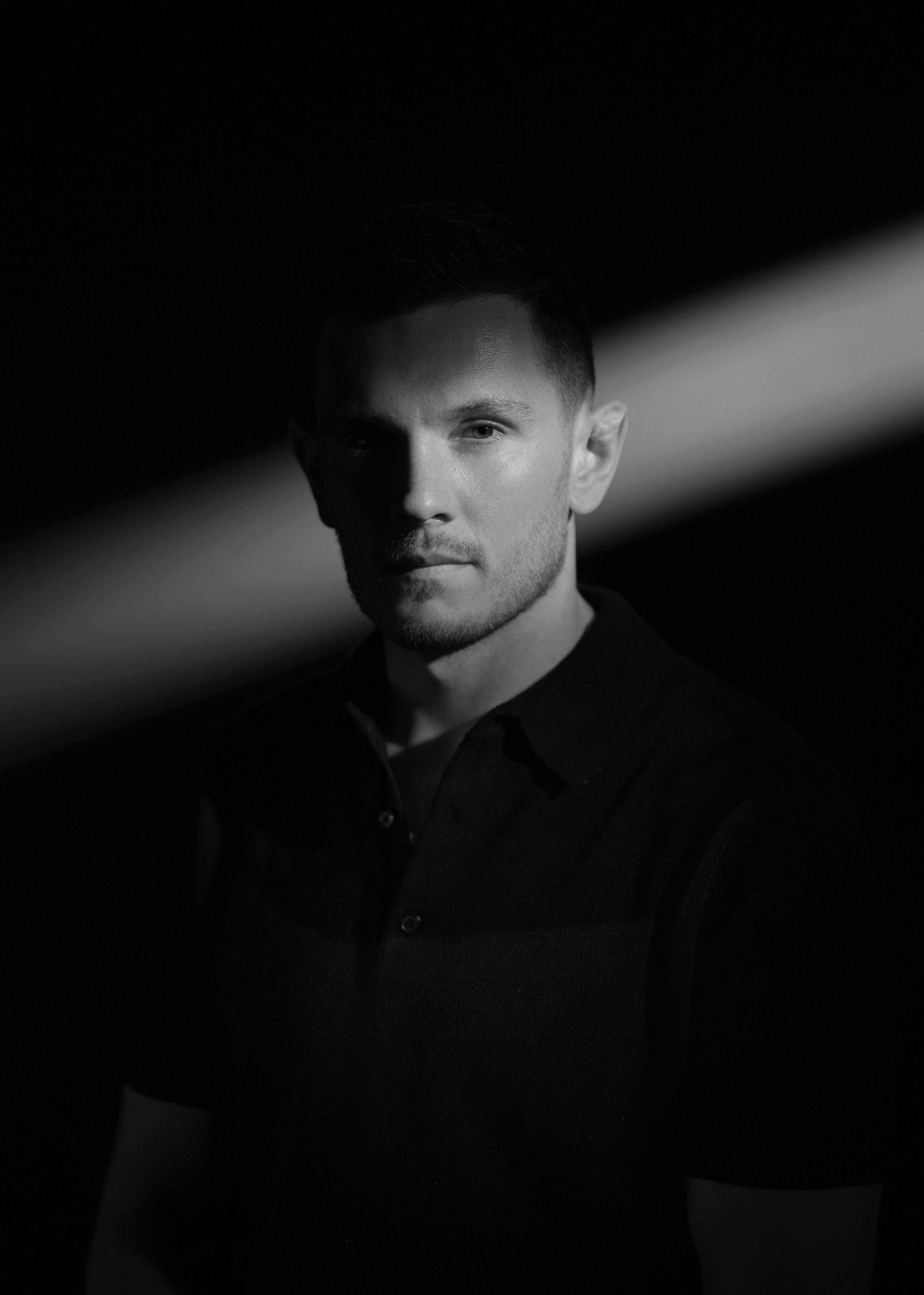 Black and white portrait of a young man with short hair wearing a dark collared shirt, looking directly at the camera with half of his face illuminated.